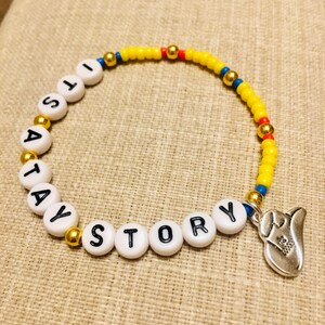 May include: A beaded bracelet featuring yellow, blue, and red seed beads, gold-toned metallic spacer beads, and white circular beads with black letters spelling out ITS A TAY STORY. A silver-toned metal charm shaped like a cowboy boot hangs from the end.