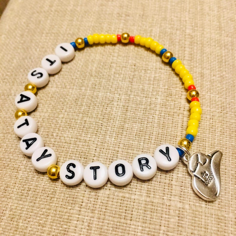 May include: A beaded bracelet featuring yellow, blue, and red seed beads, gold-toned metallic spacer beads, and white circular beads with black letters spelling out ITS A TAY STORY. A silver-toned metal charm shaped like a cowboy boot hangs from the end.