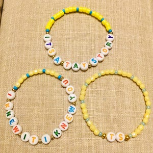 May include: Three beaded bracelets featuring yellow, blue, and gold-toned beads. One bracelet displays the text ITS A TAY STORY, another reads I KNEW IT, and the third features the letters TS, all using white letter beads with colorful text.