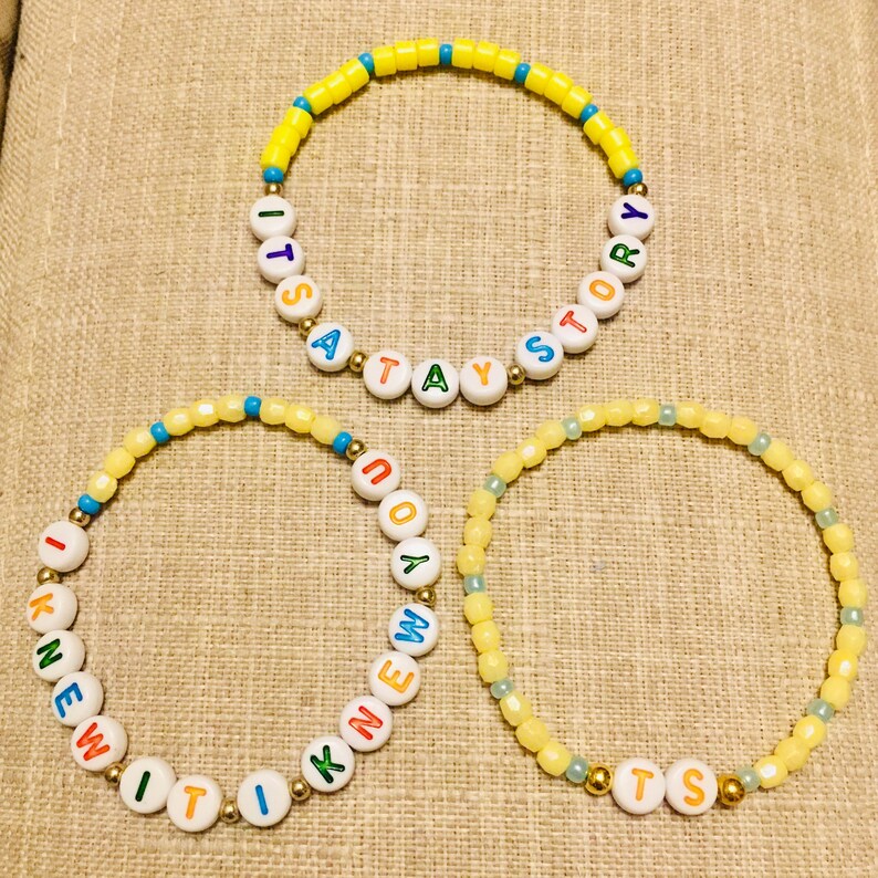 May include: Three beaded bracelets featuring yellow, blue, and gold-toned beads. One bracelet displays the text ITS A TAY STORY, another reads I KNEW IT, and the third features the letters TS, all using white letter beads with colorful text.
