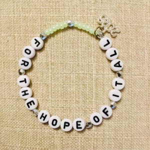 May include: A bracelet made of white letter beads spelling "FOR THE HOPE OF IT ALL". The bracelet has a light green beaded section and a silver charm with the year "2022".