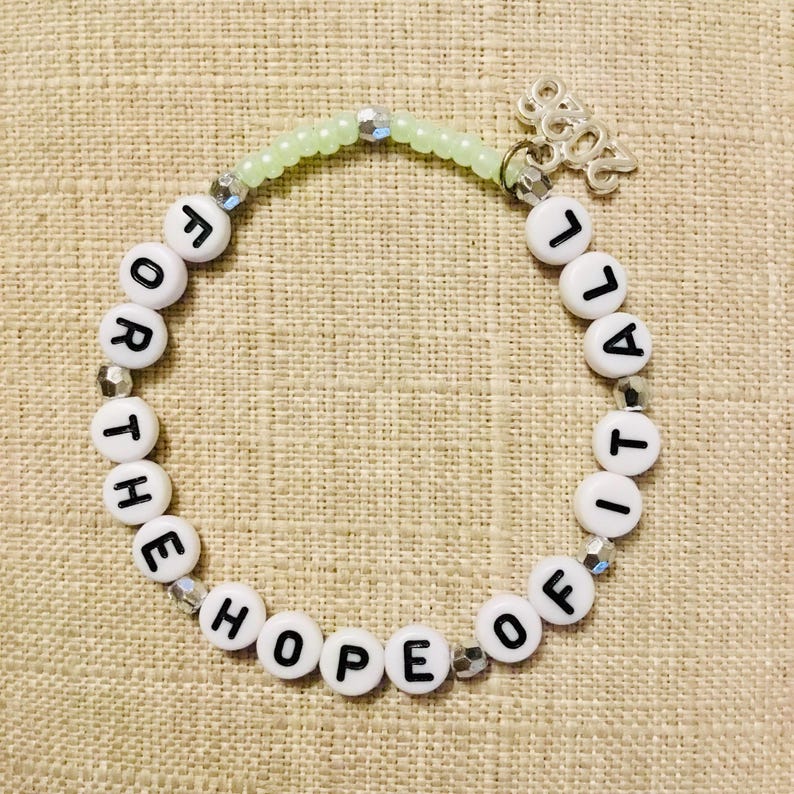 May include: A bracelet made of white letter beads spelling "FOR THE HOPE OF IT ALL". The bracelet has a light green beaded section and a silver charm with the year "2022".