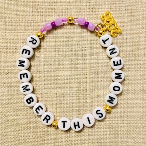 May include: A beaded bracelet with white letter beads spelling "REMEMBER THIS MOMENT". The bracelet features gold and purple beads, and a gold-coloured charm. The bracelet is on a tan surface.