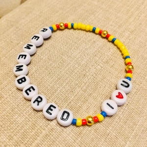 May include: A circular beaded bracelet featuring white letter beads spelling out REMEMBERED U, a red heart bead, small yellow, blue, and red seed beads, and metallic gold-toned spacer beads arranged in a repeating pattern along the elastic cord.