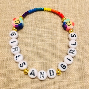 Op de afbeelding: Een kralenarmband met een regenboogkleurige boog van kleine kraaltjes, twee bloemvormige kralen met lachende gezichtjes en witte ronde kralen met zwarte letters die GIRLS AND GIRLS spellen, geaccentueerd door kleine goudkleurige bolvormige kralen.