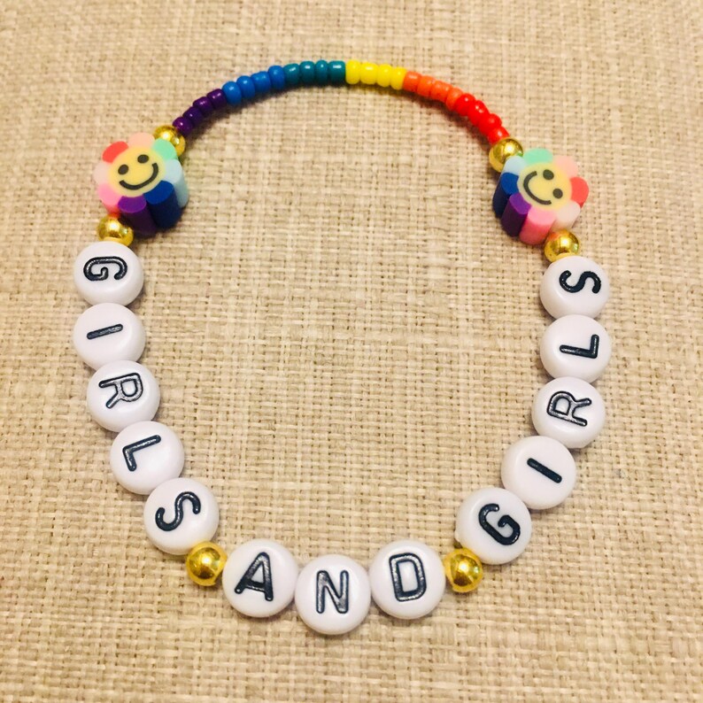 Op de afbeelding: Een kralenarmband met een regenboogkleurige boog van kleine kraaltjes, twee bloemvormige kralen met lachende gezichtjes en witte ronde kralen met zwarte letters die GIRLS AND GIRLS spellen, geaccentueerd door kleine goudkleurige bolvormige kralen.