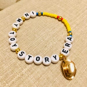 May include: A beaded bracelet featuring white round beads with black letters spelling out IN MY TOY STORY ERA, interspersed with metallic gold beads, a section of yellow seed beads, and a dangling metallic gold cowboy hat charm.