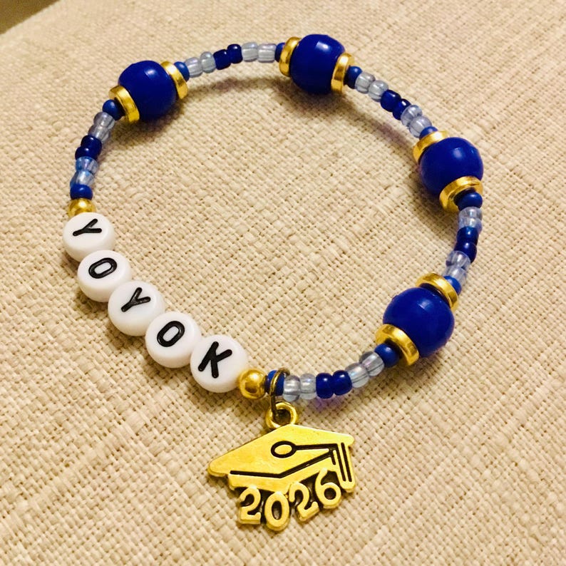 May include: A beaded bracelet with blue and gold detailing. The bracelet has white letter beads spelling "YOYOK" and a gold graduation cap charm with "2026" inscribed. The bracelet is strung with small blue and clear beads.