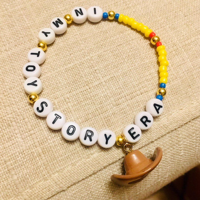 May include: A beaded bracelet featuring white letter beads spelling out IN MY TOY STORY ERA, interspersed with metallic gold beads, small yellow seed beads, and a brown cowboy hat charm dangling from the bottom of the circular jewelry piece.