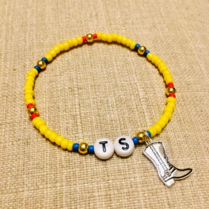May include: A beaded bracelet featuring small yellow, blue, and red beads interspersed with metallic gold spheres. Two white round beads with the black letters T and S are centered, accompanied by a silver-toned cowboy boot charm pendant.