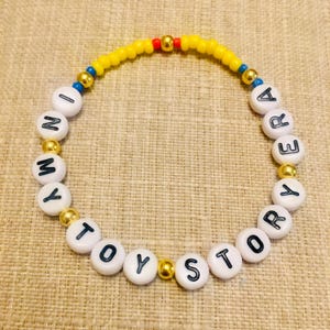 May include: A circular beaded bracelet featuring white round beads with black letters spelling out IN MY TOY STORY ERA. The design includes small yellow, red, and blue seed beads interspersed with metallic gold spherical accent beads throughout.