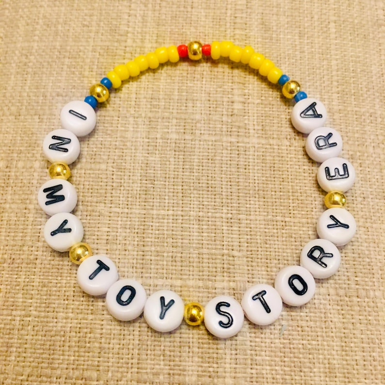 May include: A circular beaded bracelet featuring white round beads with black letters spelling out IN MY TOY STORY ERA. The design includes small yellow, red, and blue seed beads interspersed with metallic gold spherical accent beads throughout.