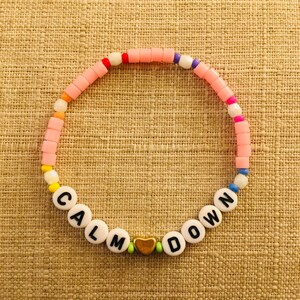 Op de afbeelding: Een cirkelvormige kralenarmband met een herhalend patroon van kleine, kleurrijke plastic kralen in tinten roze, oranje, geel, wit, blauw en paars. De armband bevat witte letterkralen die CALM DOWN spellen en een gouden hartvormige kraal.