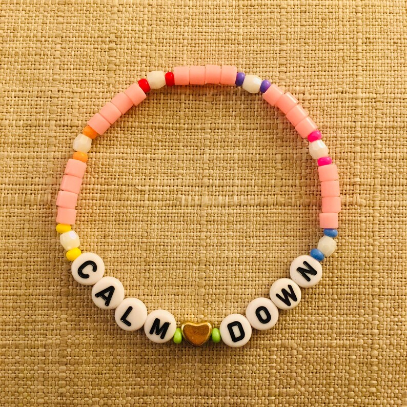 Op de afbeelding: Een cirkelvormige kralenarmband met een herhalend patroon van kleine, kleurrijke plastic kralen in tinten roze, oranje, geel, wit, blauw en paars. De armband bevat witte letterkralen die CALM DOWN spellen en een gouden hartvormige kraal.