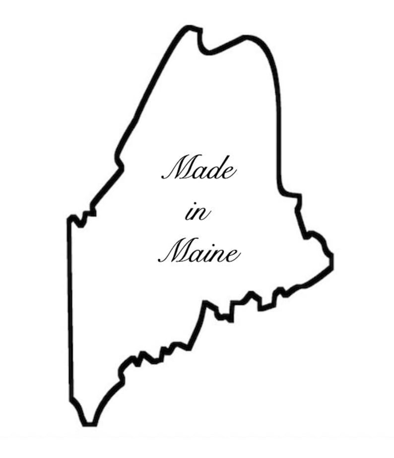 May include: A black outline of the state of Maine on a white background. The words "Made in Maine" are written in a cursive font inside the state outline.