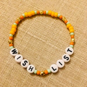 May include: A beaded bracelet with the words "WISH LIST" spelled out in white letter beads. The bracelet features orange, gold, and light green beads. The bracelet is on a tan surface.