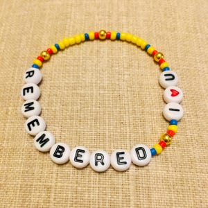 May include: A circular beaded bracelet featuring white alphabet beads spelling out REMEMBERED and UAI with a red heart bead. The design includes small yellow, red, and blue seed beads interspersed with metallic gold spherical beads throughout the strand.
