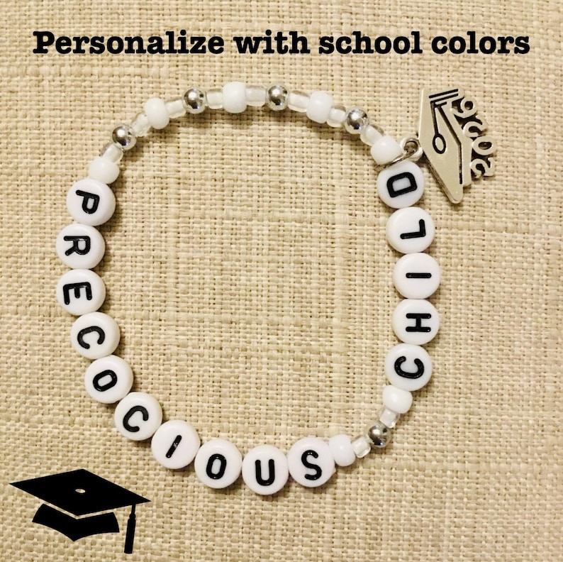May include: White beaded bracelet with the words "PRECIOUS CHILD" spelled out in black letters. The bracelet has a graduation cap charm and the text "Personalize with school colors".