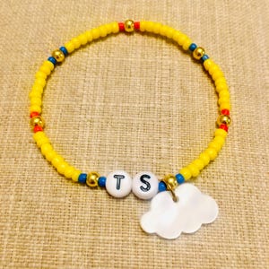 May include: A beaded bracelet featuring a circular strand of small yellow beads interspersed with occasional blue, red, and metallic gold beads. Two white spherical beads with the black letters T and S are centered, accompanied by a white cloud charm.