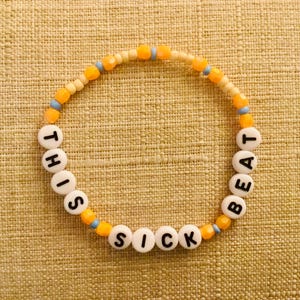 May include: A beaded bracelet with white letter beads spelling out "This Beat Is Sick" in black lettering. The beads are strung on a white and orange string.