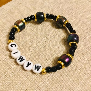 May include: A beaded bracelet with black and iridescent beads, gold accents, and white letter beads spelling "CIWYW". The bracelet features a mix of round and cylindrical beads, creating a unique texture and design. The bracelet is on a neutral-toned surface.