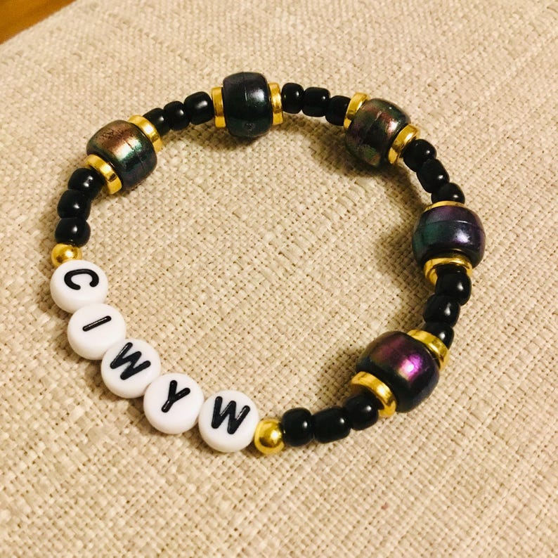 May include: A beaded bracelet with black and iridescent beads, gold accents, and white letter beads spelling "CIWYW". The bracelet features a mix of round and cylindrical beads, creating a unique texture and design. The bracelet is on a neutral-toned surface.