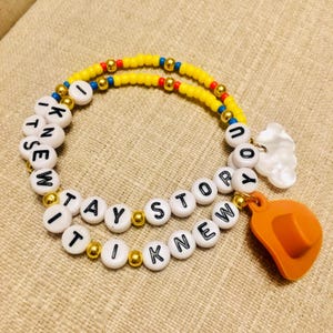 May include: A double-strand beaded bracelet featuring yellow, blue, and red seed beads interspersed with gold-tone metallic beads. White letter beads spell out the phrase I KNEW IT TAY STORY, accented by a white cloud charm and an orange cowboy hat charm.