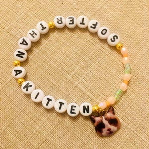 May include: A beaded bracelet with white letter beads spelling "ANOTHER SOFTER KITTEN." The bracelet features gold-colored accent beads and a charm with a cat face. The beads are strung on an elastic cord.