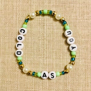 May include: A beaded bracelet with the words "COLD AS YOU" spelled out in white letter beads. The bracelet features gold, green, teal, and pearl-colored beads. The bracelet is on a neutral-colored surface.