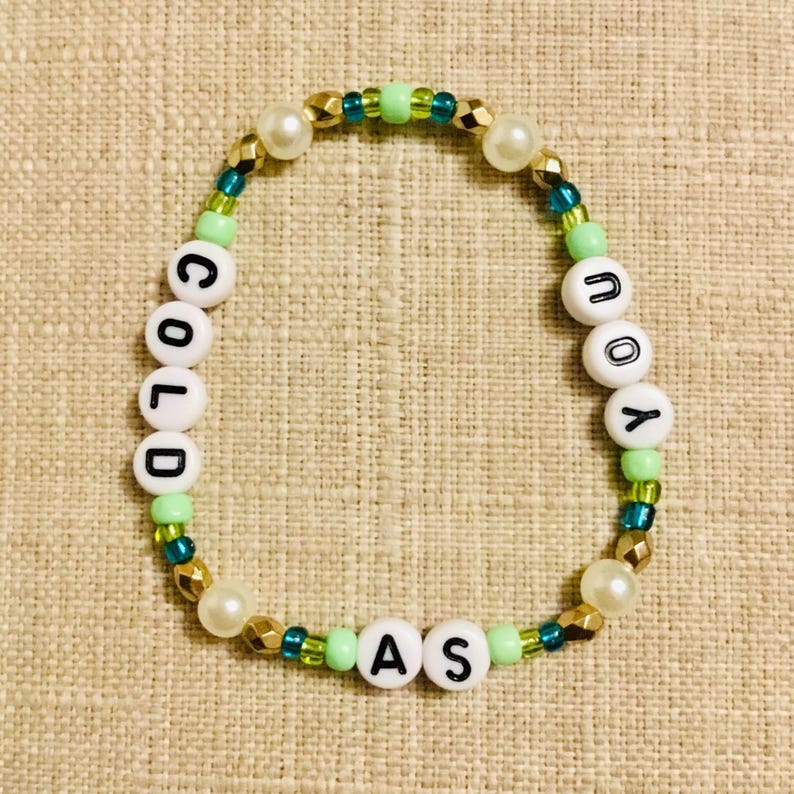 May include: A beaded bracelet with the words "COLD AS YOU" spelled out in white letter beads. The bracelet features gold, green, teal, and pearl-colored beads. The bracelet is on a neutral-colored surface.