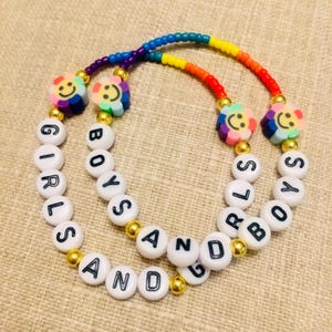 Op de afbeelding: Twee kralenarmbanden met een regenboogkleurige streng van kleine kralen, goudkleurige tussenkralen en witte letterkralen die GIRLS AND BOYS spellen. Elke armband bevat twee veelkleurige bloemvormige kralen met een smiley-gezichtje.