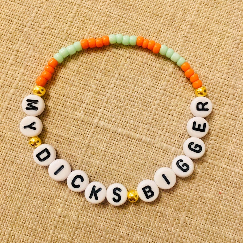 May include: A beaded bracelet with the text "MY DICKS BIGGER" spelled out in black letters on white beads. The bracelet features orange and green beads, with gold-colored beads separating the words.
