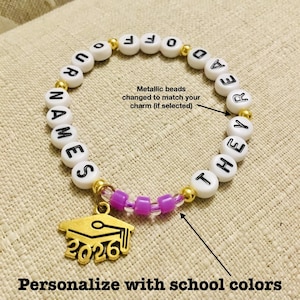 Puede incluir: Una pulsera de cuentas blancas con cuentas de letras negras que deletrean "NAMES THEYRE OFF". Cuenta con un dije de gorro de graduaci&oacute;n dorado con "2026" y cuentas moradas. El texto "Personaliza con los colores de la escuela" est&aacute; en la parte inferior.