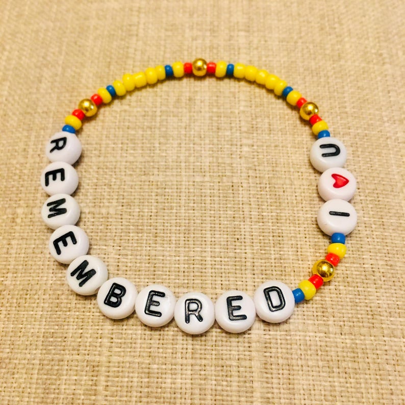 May include: A circular beaded bracelet featuring white alphabet beads spelling out REMEMBERED and UAI with a red heart bead. The design includes small yellow, red, and blue seed beads interspersed with metallic gold spherical beads throughout the strand.
