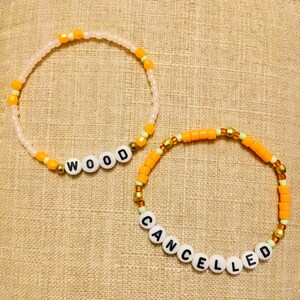 May include: Two beaded bracelets. One bracelet spells "WOOD" in white letter beads, with orange and gold accents. The other bracelet spells "CANCELLED" in white letter beads, with orange and gold accents. Both bracelets are on a tan surface.