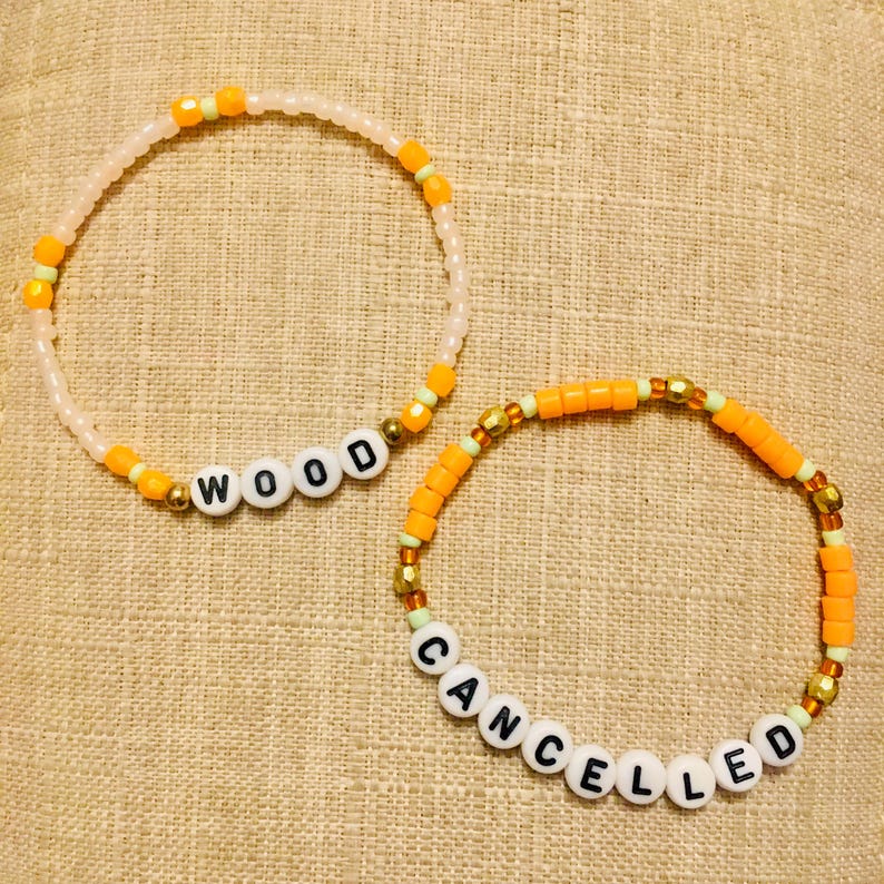 May include: Two beaded bracelets. One bracelet spells "WOOD" in white letter beads, with orange and gold accents. The other bracelet spells "CANCELLED" in white letter beads, with orange and gold accents. Both bracelets are on a tan surface.