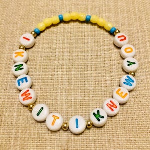 May include: A circular beaded bracelet featuring white round beads with colorful letters spelling I KNEW IT I KNEW YOU, interspersed with small metallic gold beads, and a top section of yellow and blue faceted beads arranged in a repeating pattern.
