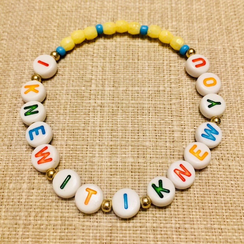 May include: A circular beaded bracelet featuring white round beads with colorful letters spelling I KNEW IT I KNEW YOU, interspersed with small metallic gold beads, and a top section of yellow and blue faceted beads arranged in a repeating pattern.