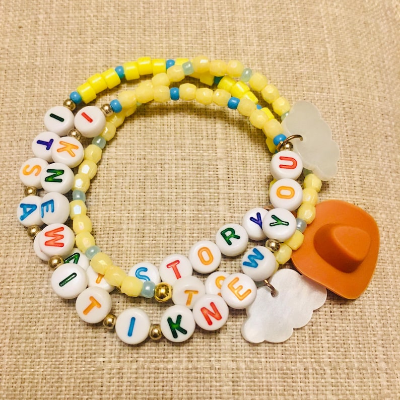 May include: A beaded bracelet featuring yellow, blue, and gold-toned beads interspersed with white letter beads spelling out "ITS A NEW STORY YOU KNEW IT". The bracelet includes a brown cowboy hat charm and two white cloud-shaped charms.