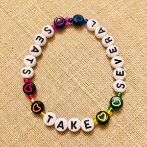 Op de afbeelding: Een ronde kralenarmband met witte alfabetkralen die de tekst TAKE SEVERAL SEATS vormen. Het ontwerp bevat zwarte kralen met gekleurde hartomlijningen in roze, blauw, oranje, geel en groen, afgewisseld met kleine metalen kralen.