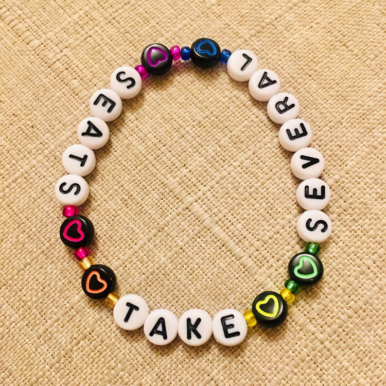 Op de afbeelding: Een ronde kralenarmband met witte alfabetkralen die de tekst TAKE SEVERAL SEATS vormen. Het ontwerp bevat zwarte kralen met gekleurde hartomlijningen in roze, blauw, oranje, geel en groen, afgewisseld met kleine metalen kralen.