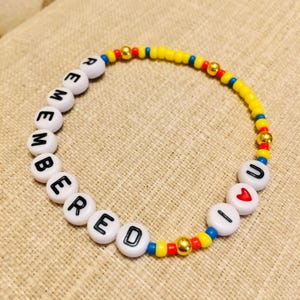 May include: A circular beaded bracelet featuring white letter beads spelling out REMEMBERED U, a red heart bead, small yellow, blue, and red seed beads, and metallic gold-toned spacer beads arranged in a repeating pattern along the elastic cord.