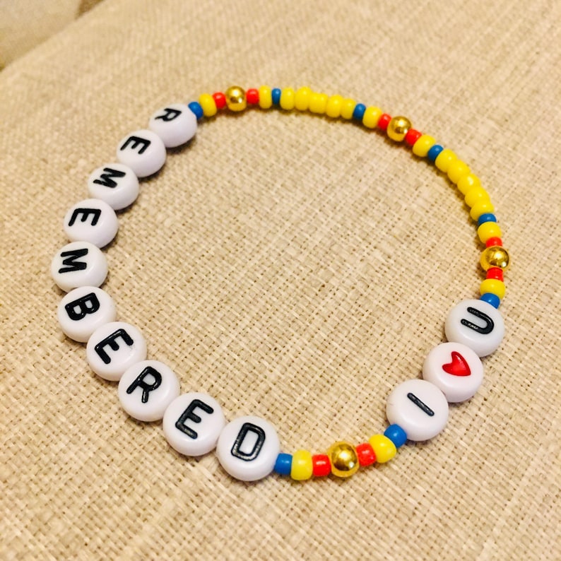 May include: A circular beaded bracelet featuring white letter beads spelling out REMEMBERED U, a red heart bead, small yellow, blue, and red seed beads, and metallic gold-toned spacer beads arranged in a repeating pattern along the elastic cord.