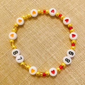 May include: A beaded bracelet featuring white letter beads with the numbers 7, 8, and 6, and white heart-shaped beads with orange and red heart accents. The bracelet is strung with gold and red beads.