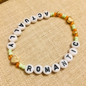 May include: A beaded bracelet with the words "ACTUALLY ROMANTIC" spelled out in black letters on white beads. The bracelet features gold heart charms and is strung with green and orange beads.