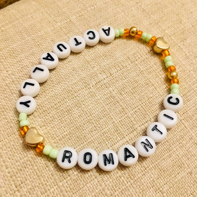May include: A beaded bracelet with the words "ACTUALLY ROMANTIC" spelled out in black letters on white beads. The bracelet features gold heart charms and is strung with green and orange beads.