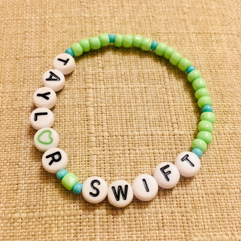 May include: A beaded bracelet with the name "TAYLOR SWIFT" spelled out in black letters on white beads. The bracelet features green and turquoise beads, with a heart symbol on one bead. The bracelet is on a tan surface.