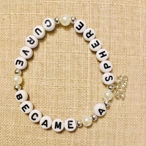 May include: White beaded bracelet with black letter beads spelling "CURE BECAME SPHERE". The bracelet includes pearl and silver accent beads, and a small silver charm with the year "2020".