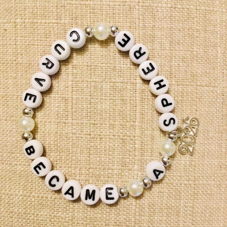 May include: White beaded bracelet with black letter beads spelling "CURE BECAME SPHERE". The bracelet includes pearl and silver accent beads, and a small silver charm with the year "2020".