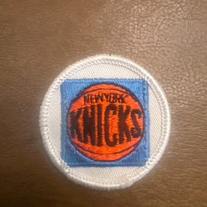 May include: A circular embroidered patch featuring a white border and a light blue square background. Centered on the square is an orange basketball graphic with the text NEW YORK KNICKS embroidered in dark blue lettering across the centre.